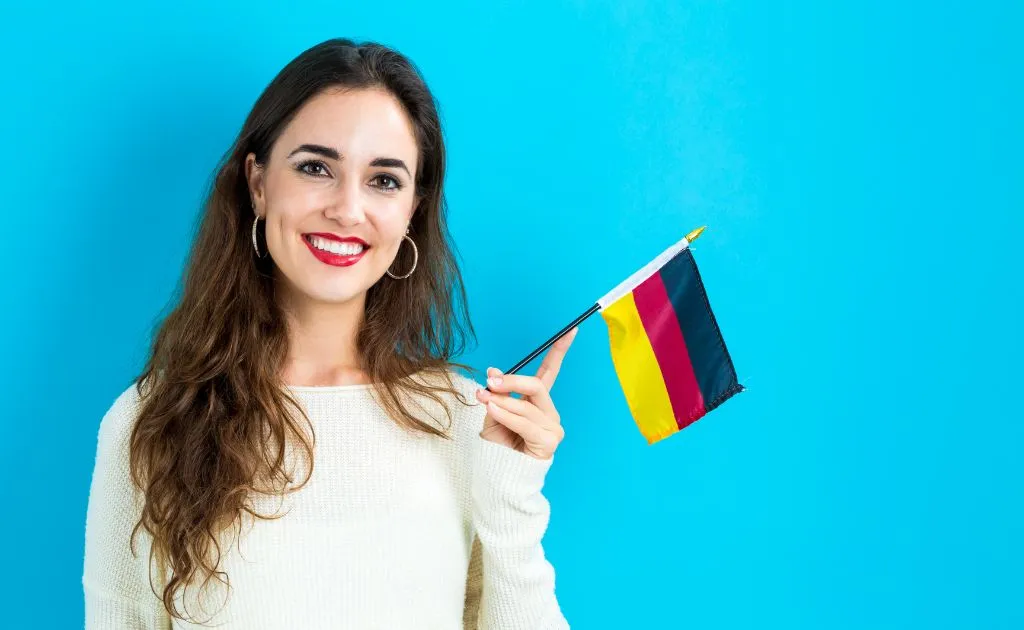 Ragazza sorridente che tiene in mano una bandiera tedesca su sfondo azzurro, rappresentando i corsi di lingua disponibili su Preply