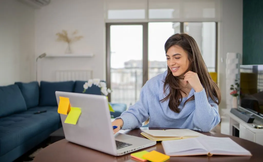 Studentessa sorridente studia al laptop con quaderno aperto, ripetizioni online da casa