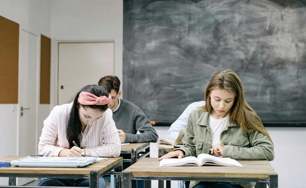 Studenti concentrati che leggono e scrivono ai banchi di scuola con la lavagna sullo sfondo